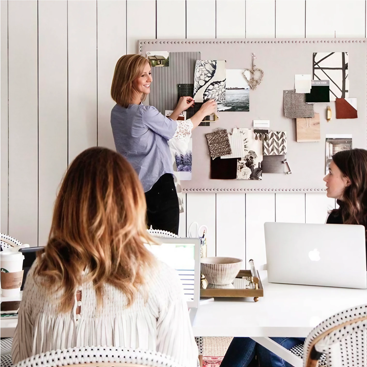 Three interior designers working in a bright studio; one pins fabric and wallpaper swatches to a large pinboard while two colleagues review the scheme from a table of laptops