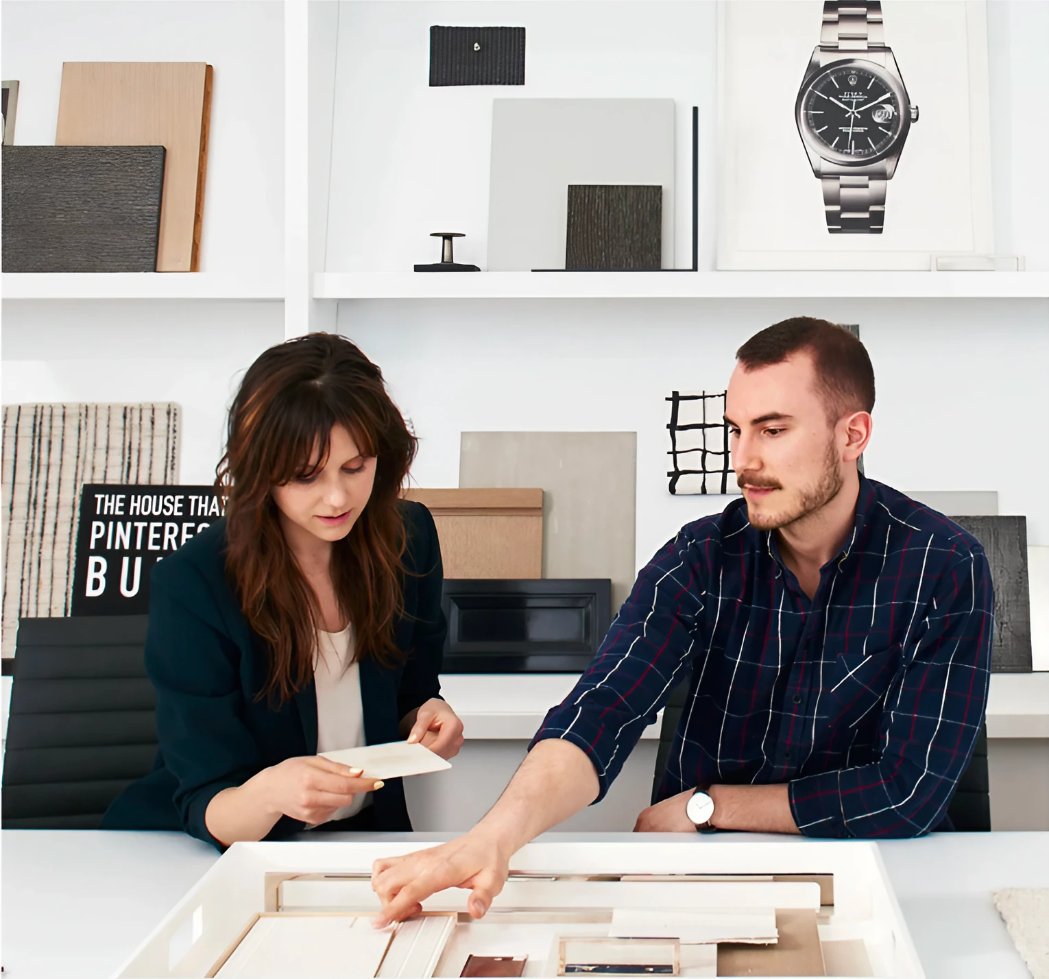 Two designers at a white table comparing paper samples over an open drawer of material swatches, with shelves of wood, fabric and stone finishes behind them